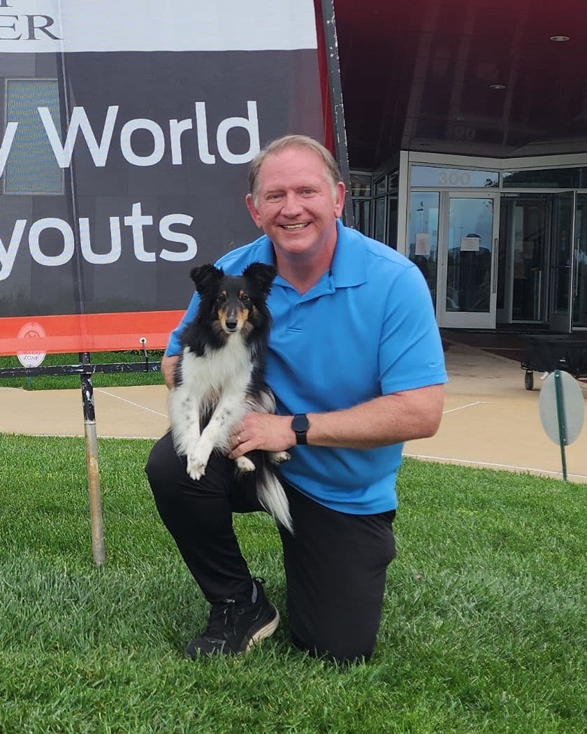 Lloyd Silver kneeling on grass, holding his Sheltie Vespa, in front of an AKC Agility World Team Tryouts banner